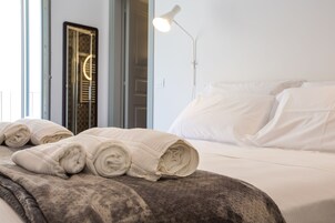 Una cama impecable con toallas enrolladas, una lámpara de noche y vista a una puerta con un espejo circular.
