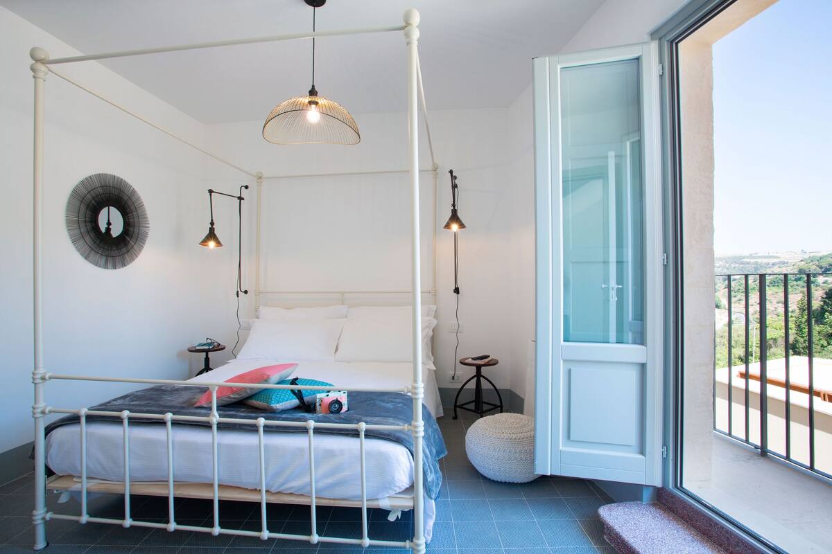 A four-poster bed with a white frame, a round mirror on the wall, and a view of the outdoors through a glass door.