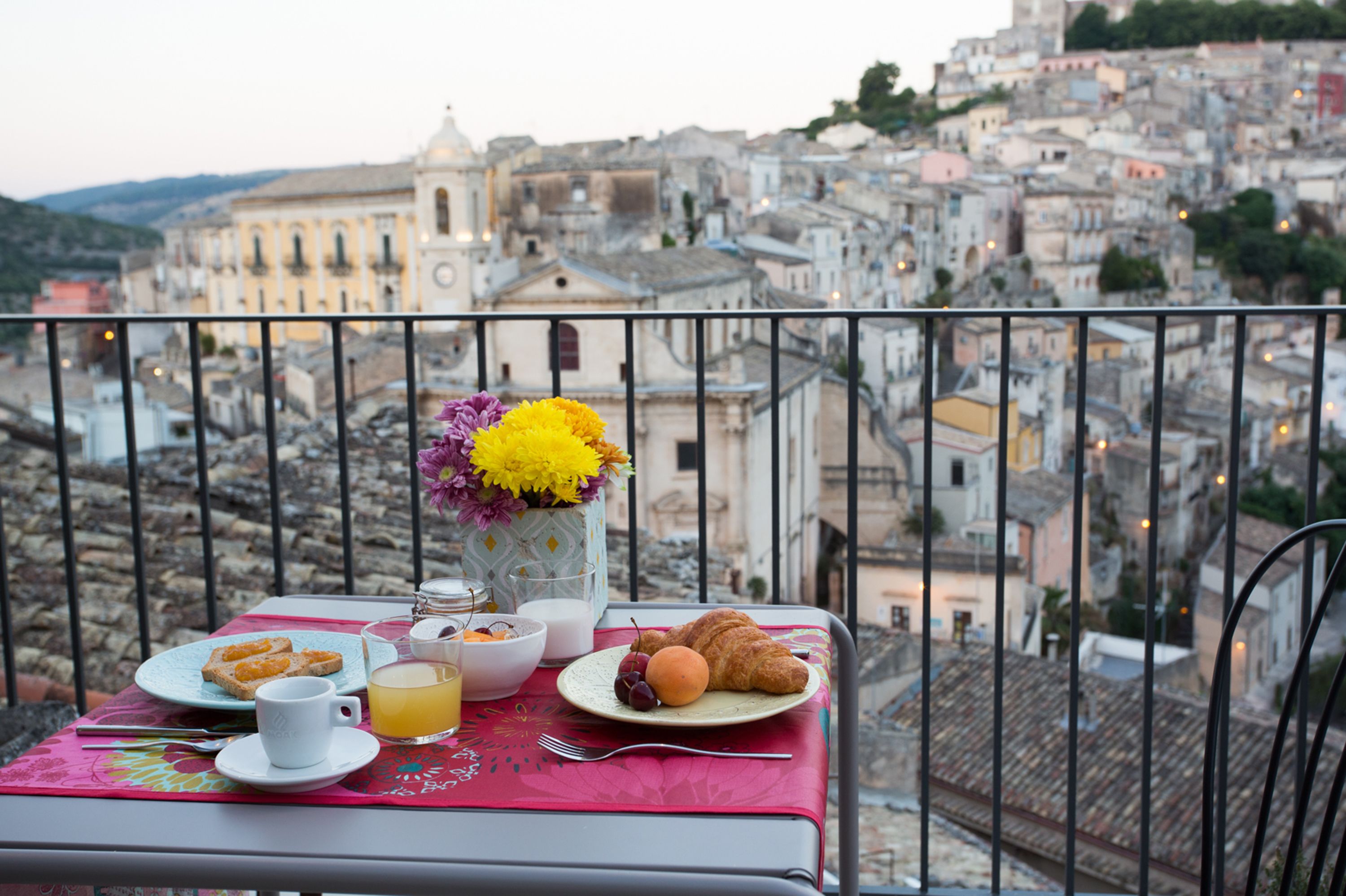 A balcony table with breakfast items, a vase of flowers, and a cityscape view.