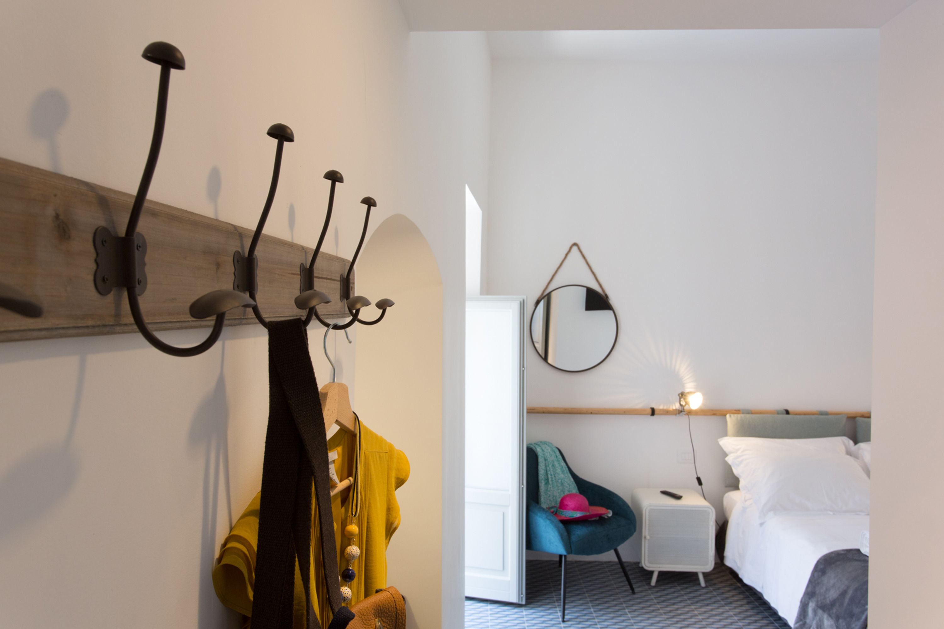 Habitación de hotel con cama, mesita de noche con lámpara, una silla y perchero con varias prendas colgadas.