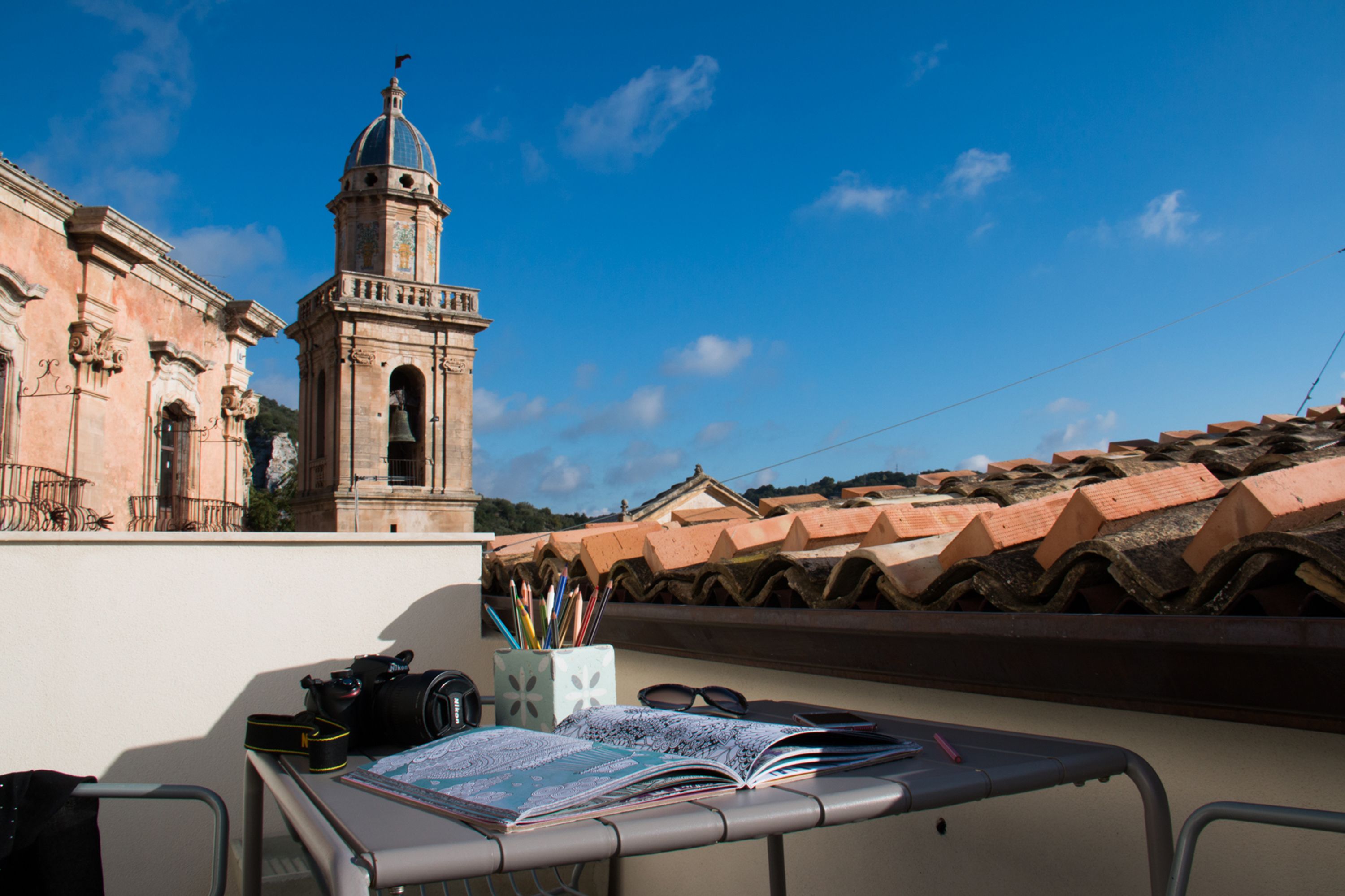 A balcony with a table set up, featuring a camera, books, and a cup of pens. A bell tower is visible in the background.