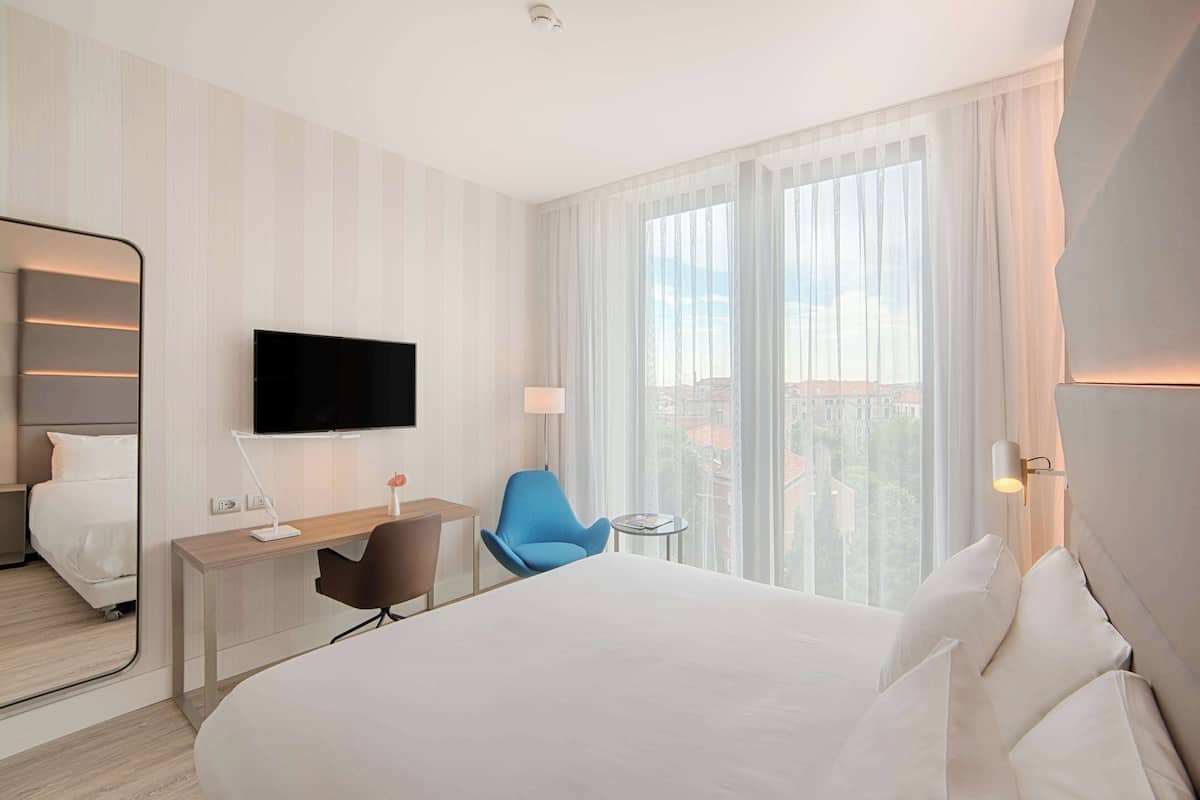 A hotel room with a bed, a desk, a chair, a TV, and a window with a view.