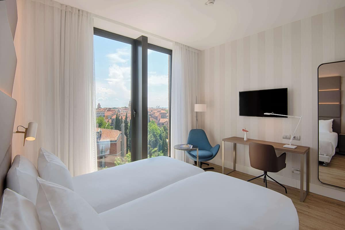 A hotel room with a large bed, a desk, a chair, a TV, and a view of the city.
