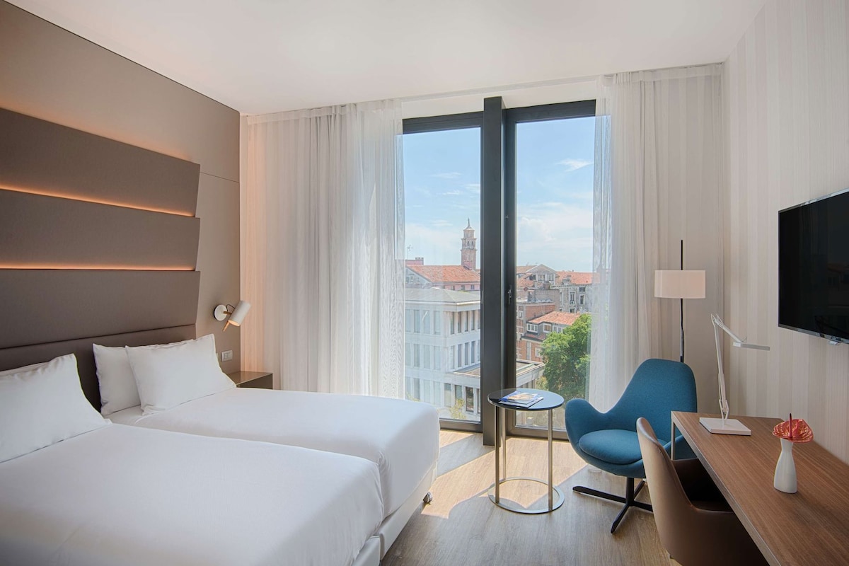 Quarto de hotel com duas camas, uma escrivaninha, uma cadeira, uma televisão e vista para a cidade.