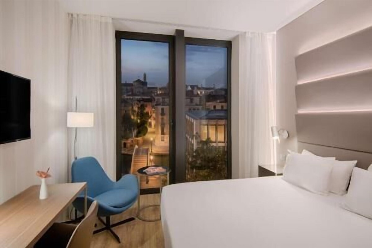 A hotel room with a bed, a desk, a chair, a TV, and a large window with a city view.