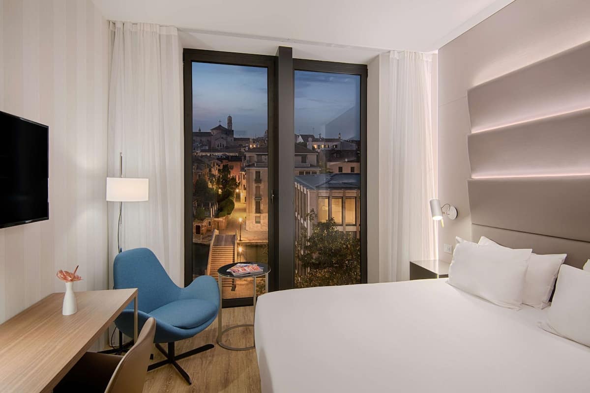 A hotel room with a bed, a desk with a chair, a TV, a lamp, and a view of a cityscape through large windows.