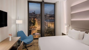 A hotel room with a bed, a desk, a chair, a TV, a lamp, a table with magazines, and a view of a cityscape through a large window. - Avani Rio Novo Venice Hotel (Venice)