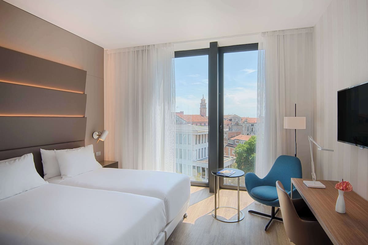 A hotel room with two beds, a desk, a chair, a TV, and a view of the city.