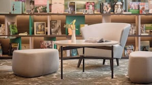 A library with a round table, two ottomans, and bookshelves filled with books. - Avani Rio Novo Venice Hotel (Venice)