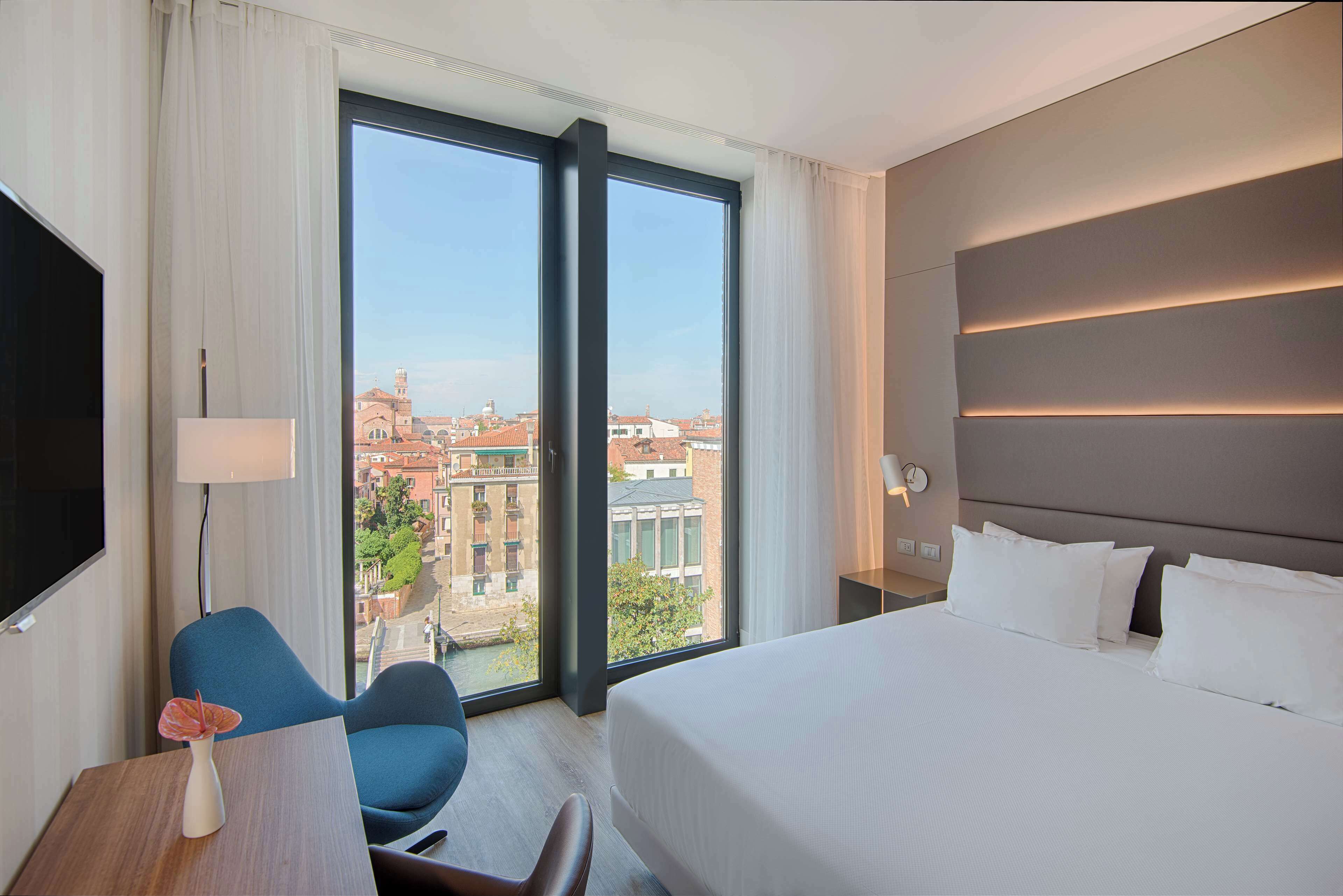 A hotel room with a large bed, a desk with a chair, a TV, and a view of the city through large windows.