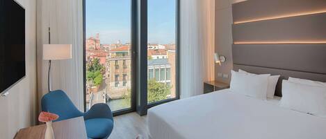 A hotel room with a large bed, a desk with a chair, a TV, and a view of the city through large windows.