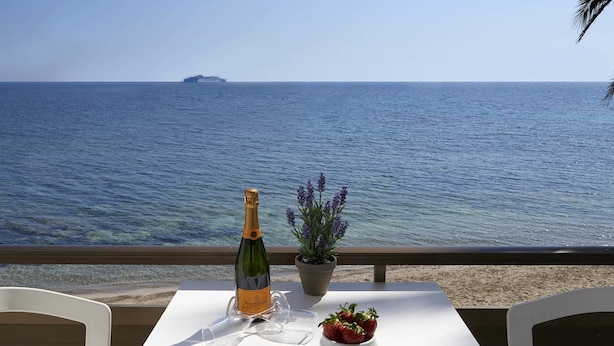 Uma mesa com uma garrafa de champanhe, um recipiente com morangos e um vaso de planta em uma varanda com vista para o mar.