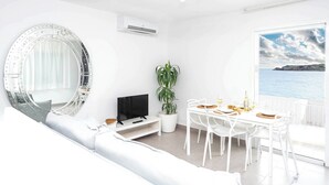 Um quarto claro e arejado, com um espelho redondo, televisão de tela plana, planta em vaso, mesa de jantar com cadeiras e vista para o mar pela janela.