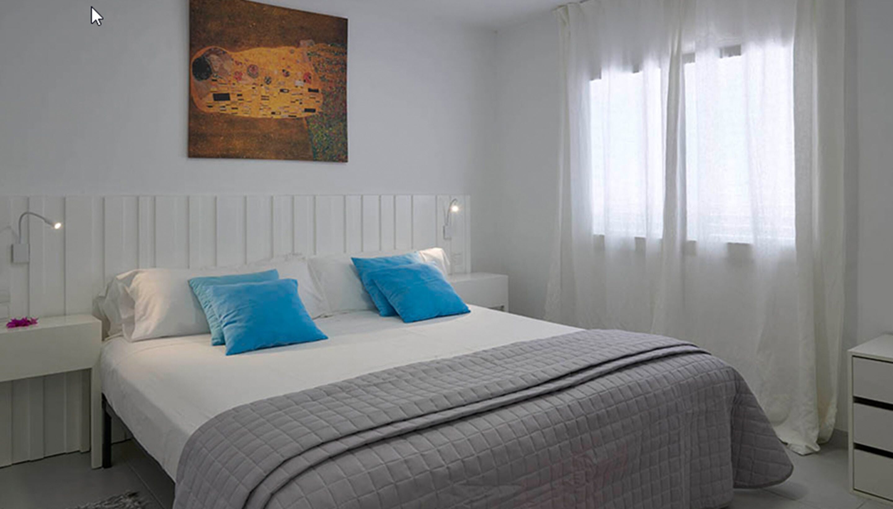 Un dormitorio con una cama grande, cabecera blanca, dos almohadas azules, colcha acolchada gris y un cuadro sobre la cama. Se ve una ventana con cortinas translúcidas.