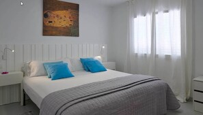Un dormitorio con una cama grande, cabecera blanca, dos almohadas azules, colcha acolchada gris y un cuadro sobre la cama. Se ve una ventana con cortinas translúcidas.