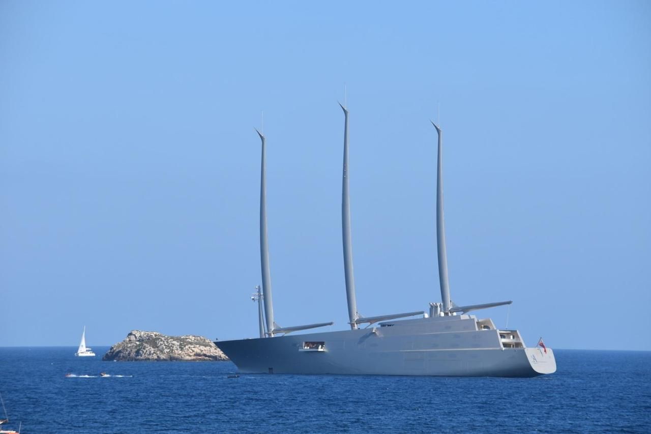 Un barco grande y moderno con varios mástiles y un diseño elegante, navegando sobre un mar tranquilo con una pequeña isla al fondo.