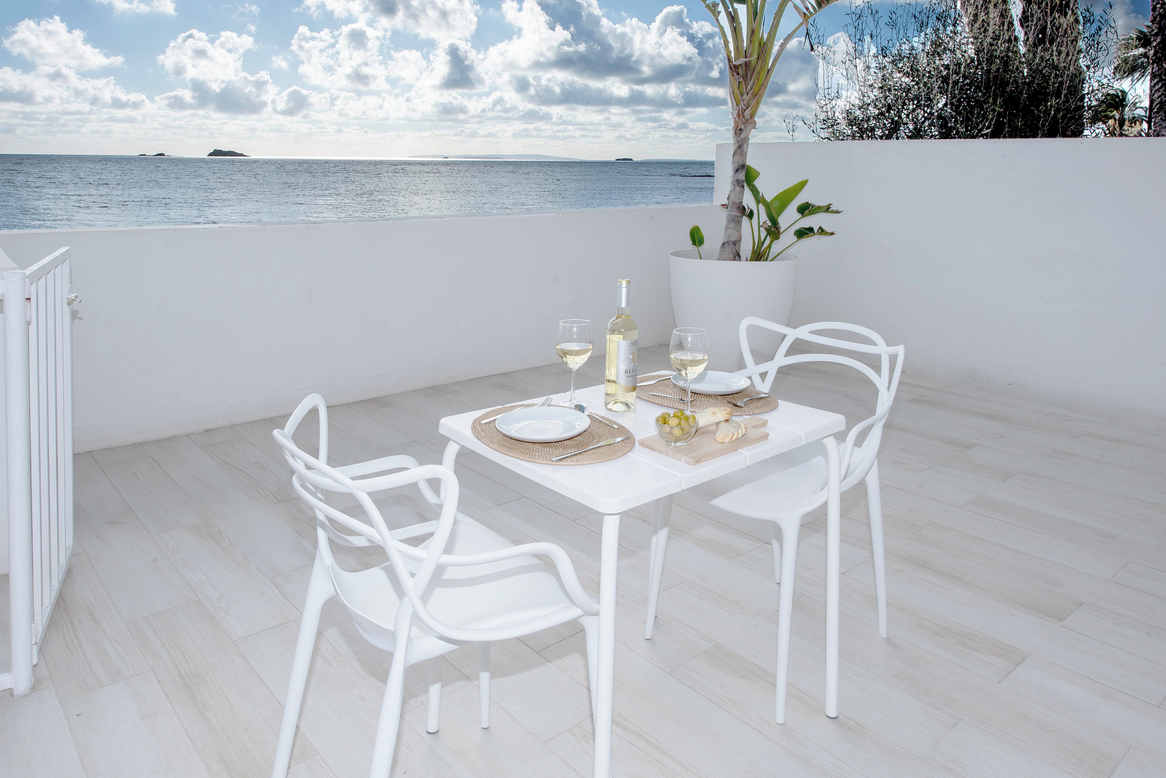 Una mesa blanca dispuesta con una botella de vino blanco, dos copas y un plato en una terraza con vistas al mar y una planta en maceta.