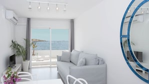 Uma sala de estar moderna com um sofá, uma mesa de jantar e cadeiras. Há uma grande janela com vista para o mar e barcos.