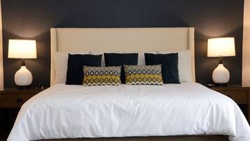 Una cama bien tendida con ropa blanca y almohadones estampados, con dos lámparas de noche a cada lado sobre mesitas a juego.