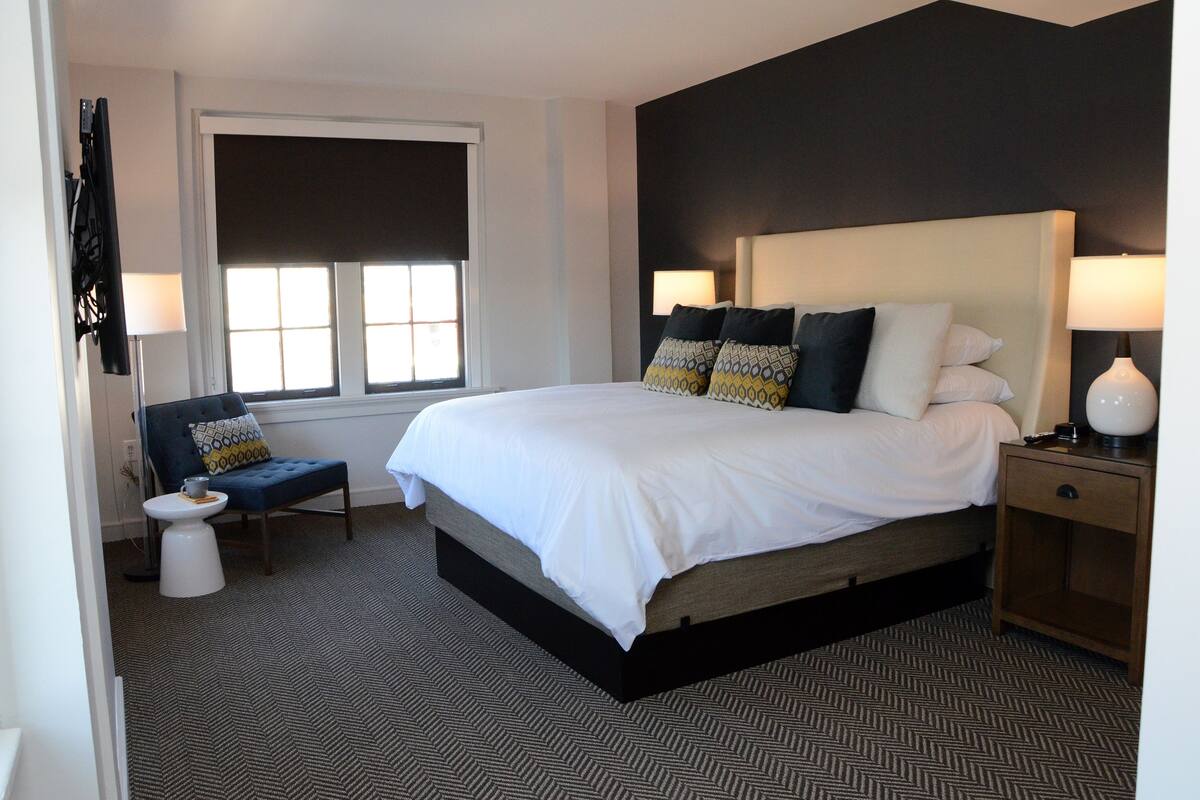 A modern hotel room with a large bed, a bedside table with a lamp, a chair, and a window with blinds.