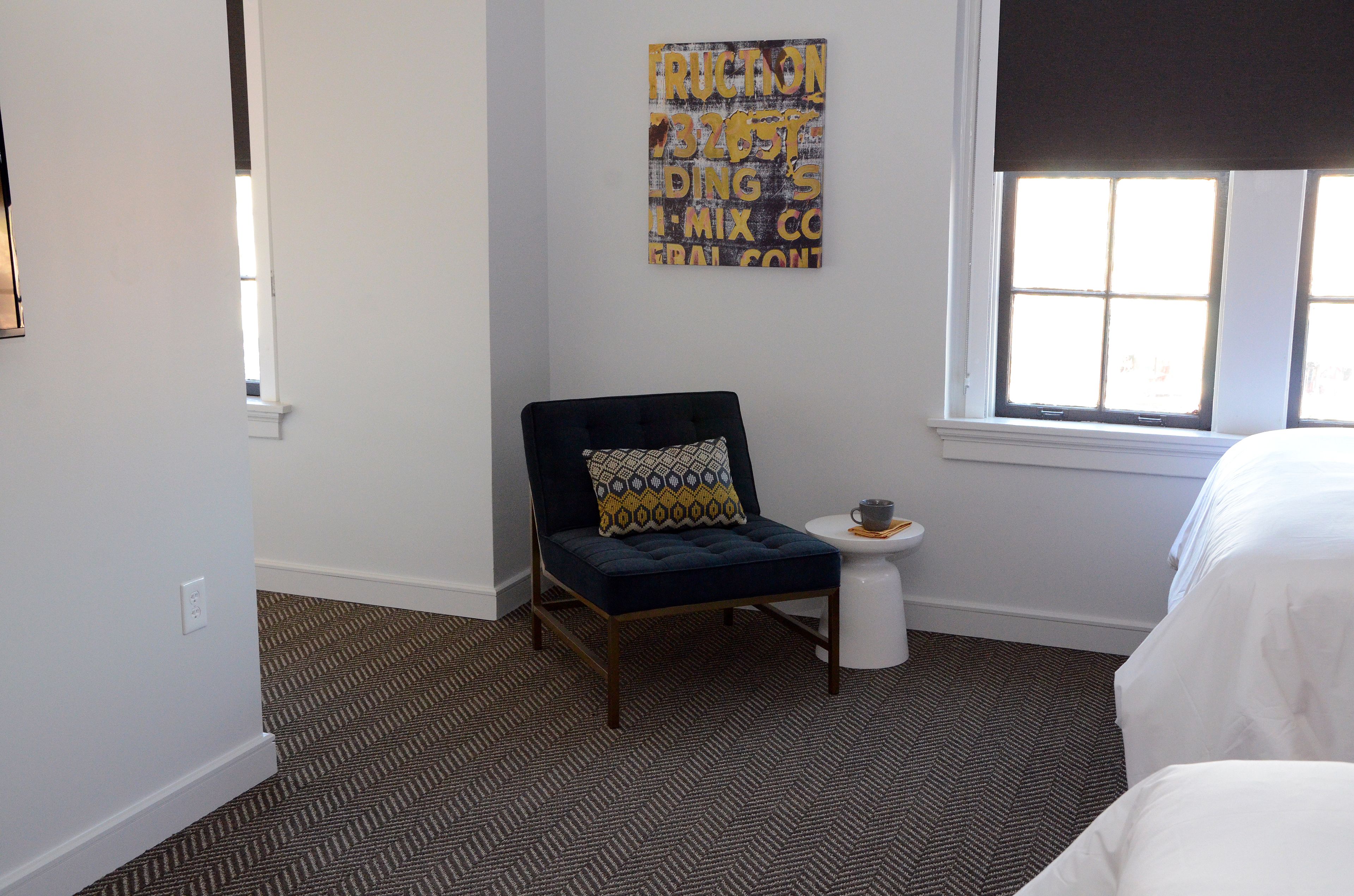 Una habitación de hotel con alfombra estampada, un sillón azul oscuro con cojín estampado, una mesita de noche blanca pequeña y una cama con ropa de cama blanca.