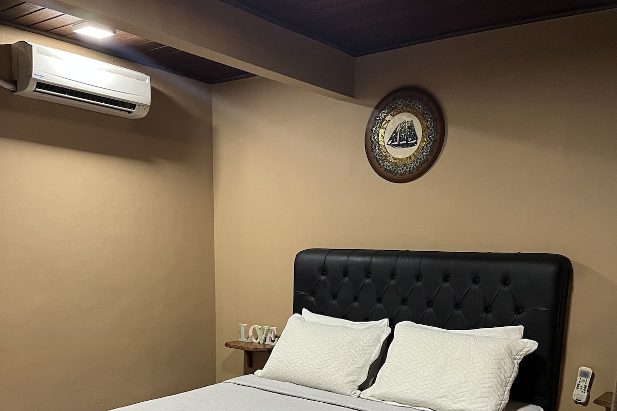 Un lit bien fait, avec une couette blanche, une tête de lit noire et un climatiseur fixé au mur.