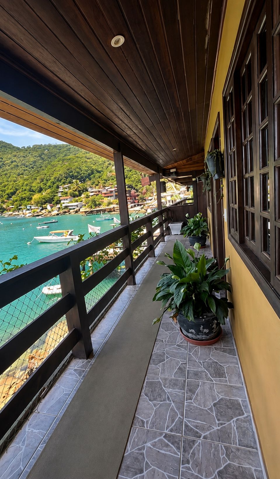 A balcony with a view of a coastal area, featuring a wooden railing, potted plants, and a stone-tiled floor.