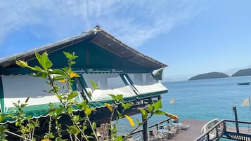 An overwater bungalows with a wooden deck and lounge chairs, overlooking a clear blue sea and distant islands.