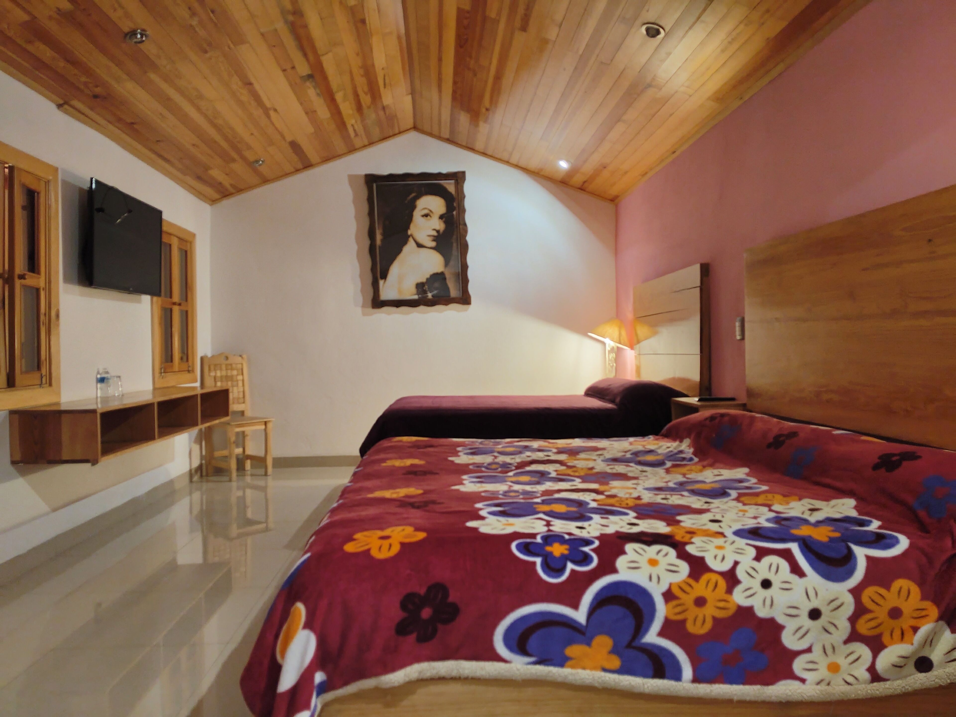 A bedroom with a wooden ceiling, a bed with a floral bedspread, a chair, a TV mounted on the wall, and a painting.