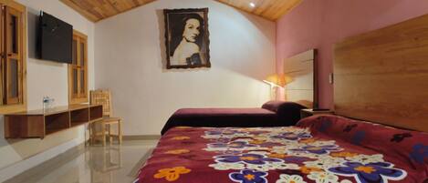 A bedroom with a wooden ceiling, a bed with a floral bedspread, a chair, a TV mounted on the wall, and a painting.