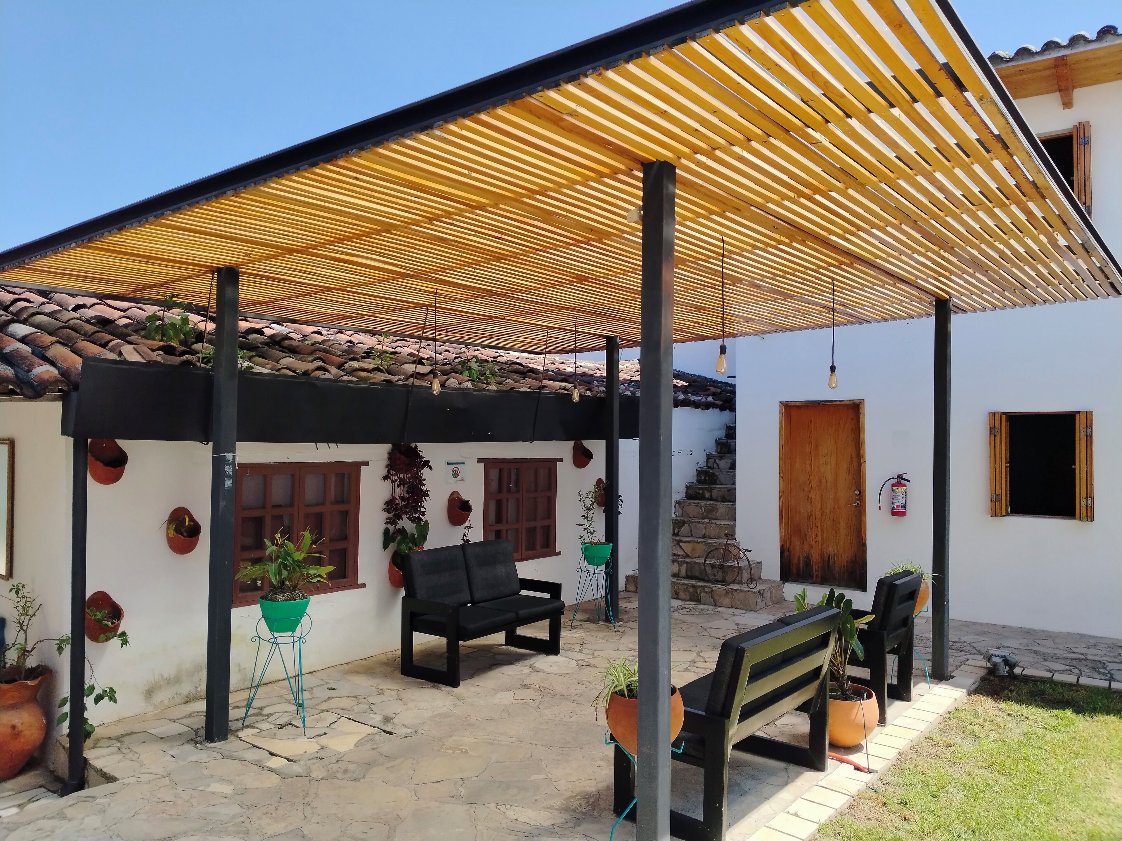 A covered patio area with a tiled roof, potted plants, a bench, and a staircase leading to a door.