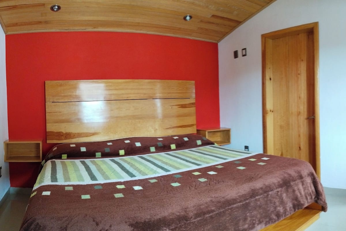 Un dormitorio con cabecera de madera, una cama con colcha estampada, una mesita de noche de madera y una pared de acento en rojo.