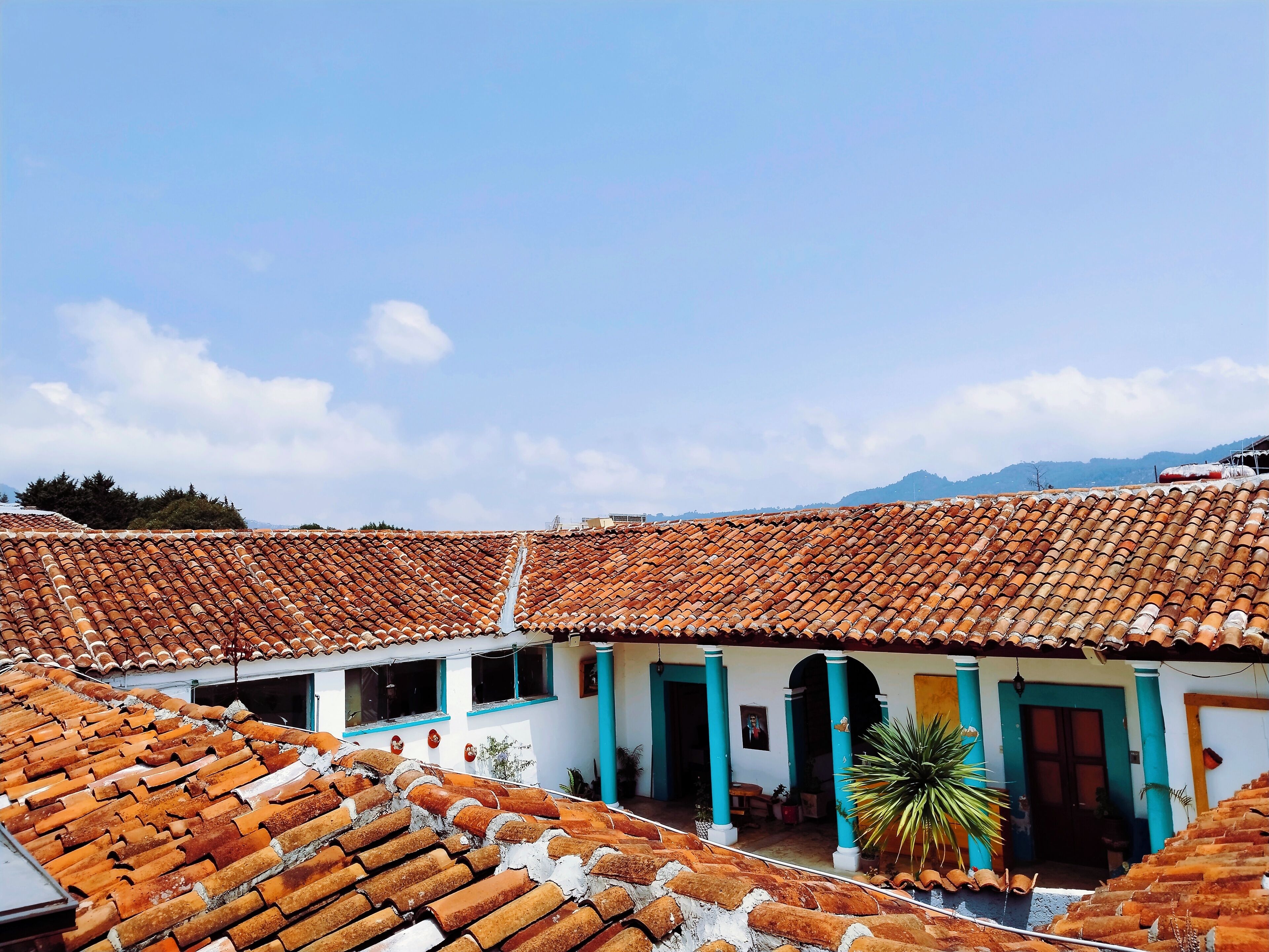 Une cour avec des toits recouverts de tuiles en terre cuite, un bâtiment blanc agrémenté de touches de bleu, et une montagne en arrière-plan.