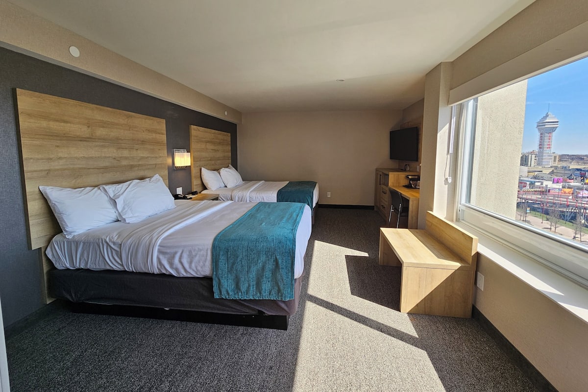 Ein Hotelzimmer mit zwei Betten, einem Schreibtisch, einem Fernseher und Blick auf eine Stadtlandschaft.