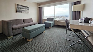 Hotelkamer met een grijze bank, een blauwe poef, een bureau met stoel en een groot raam met uitzicht op de stad.