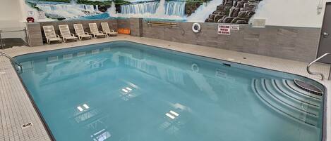 An indoor swimming pool with a mural of waterfalls, lounge chairs, and a shallow water sign.