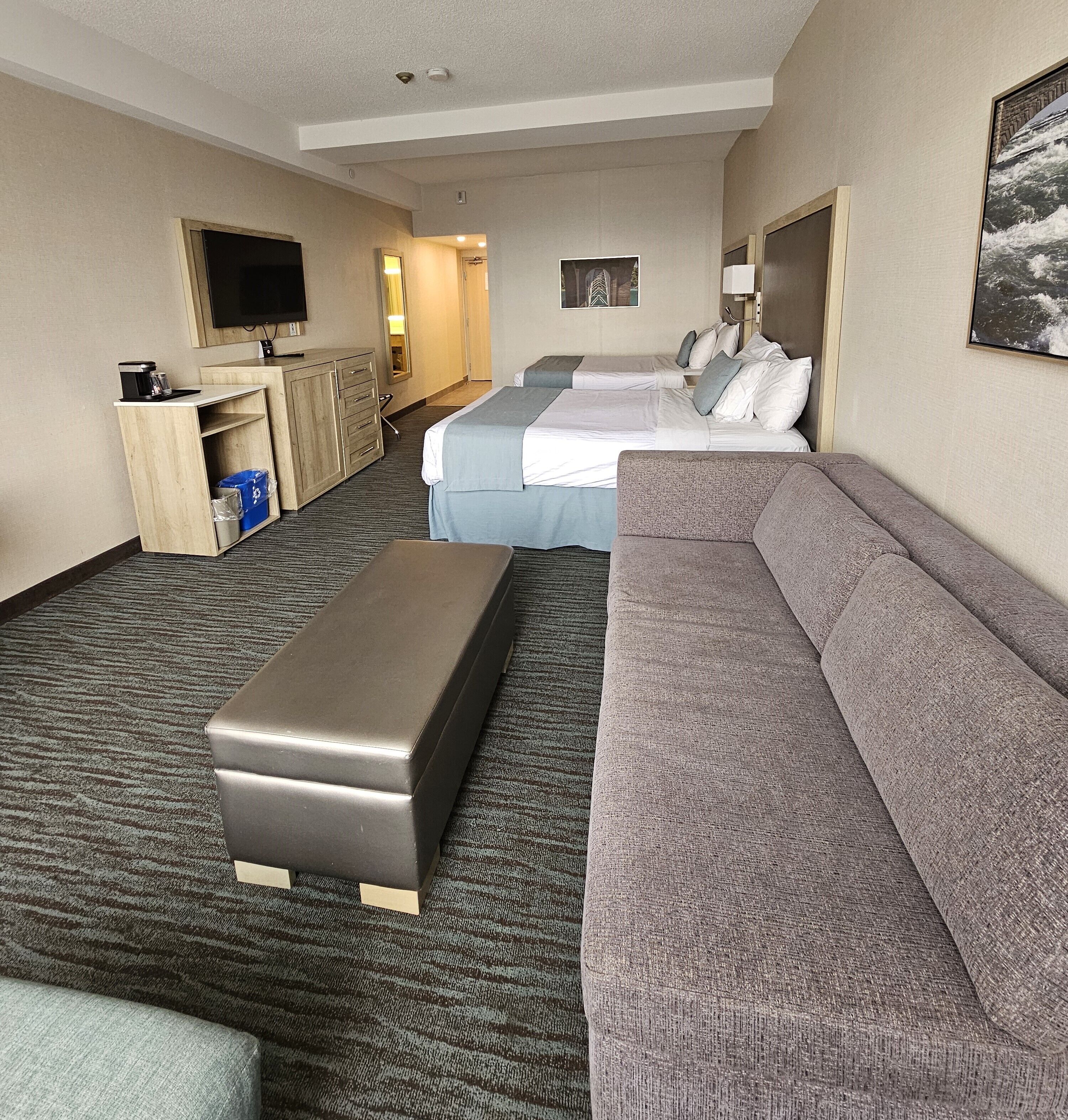 Camera d'albergo con un ampio divano grigio, un divanetto più piccolo sempre grigio, un letto con lenzuola bianche, una scrivania in legno con una televisione a schermo piatto e un pavimento rivestito di moquette.