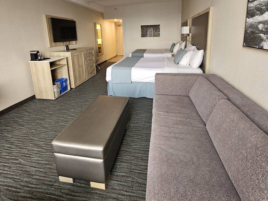 Habitación de hotel con un sofá grande de color gris, un sofá más pequeño también gris, una cama con ropa blanca, un escritorio de madera y un televisor de pantalla plana, y piso alfombrado.