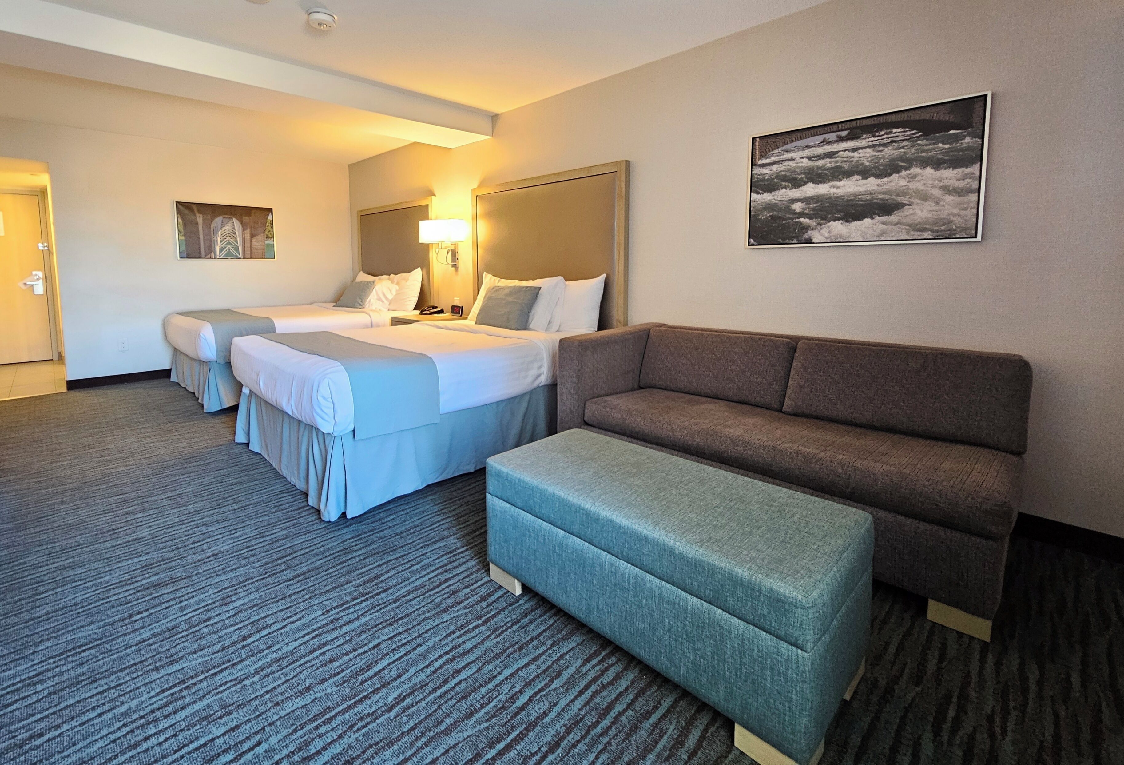 Habitación de hotel con una cama grande, un sofá y un pequeño reposapiés. Hay un cuadro en la pared y una puerta a la izquierda.