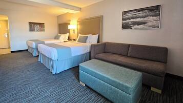 Habitación de hotel con una cama grande, un sofá y un pequeño reposapiés. Hay un cuadro en la pared y una puerta a la izquierda.