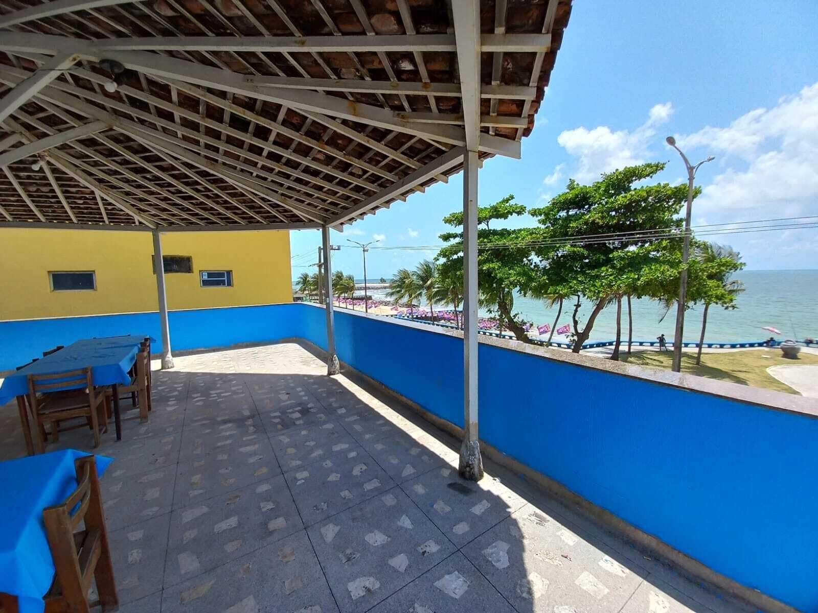 A covered patio with a view of the sea, featuring a blue and yellow building, a tiled floor, and wooden chairs.