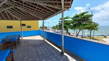 A covered patio with a view of the sea, featuring a blue and yellow building, a tiled floor, and wooden chairs.
