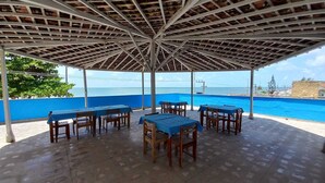 An outdoor covered patio with tables and chairs, overlooking a swimming pool and the sea. - Hotel Samburá (Olinda)