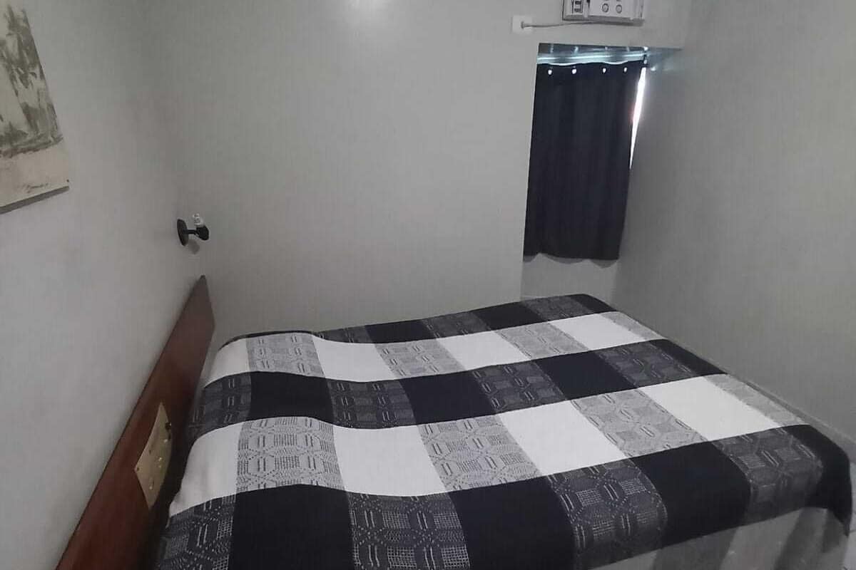 A small, single-bed room with a checkered bedspread, a wall-mounted air conditioning unit, and a curtain.