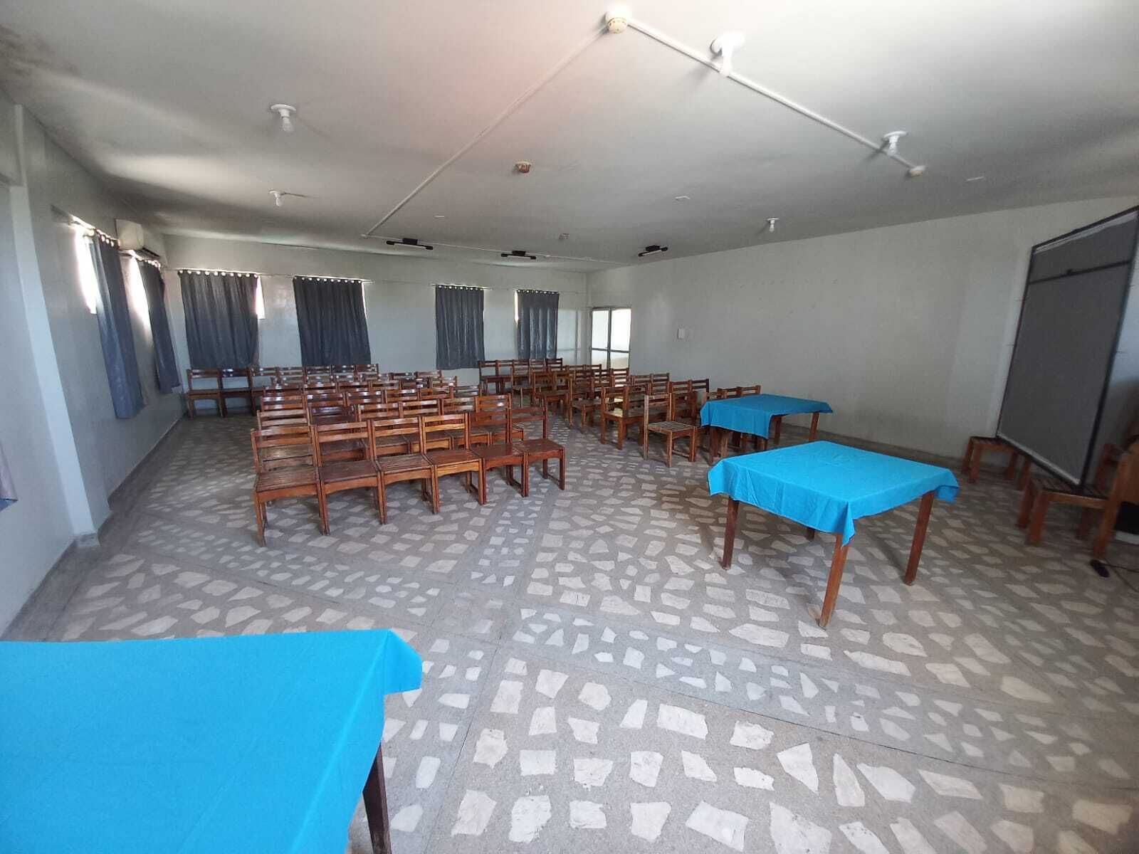 A conference room with rows of wooden chairs, blue tables, and a projector screen.