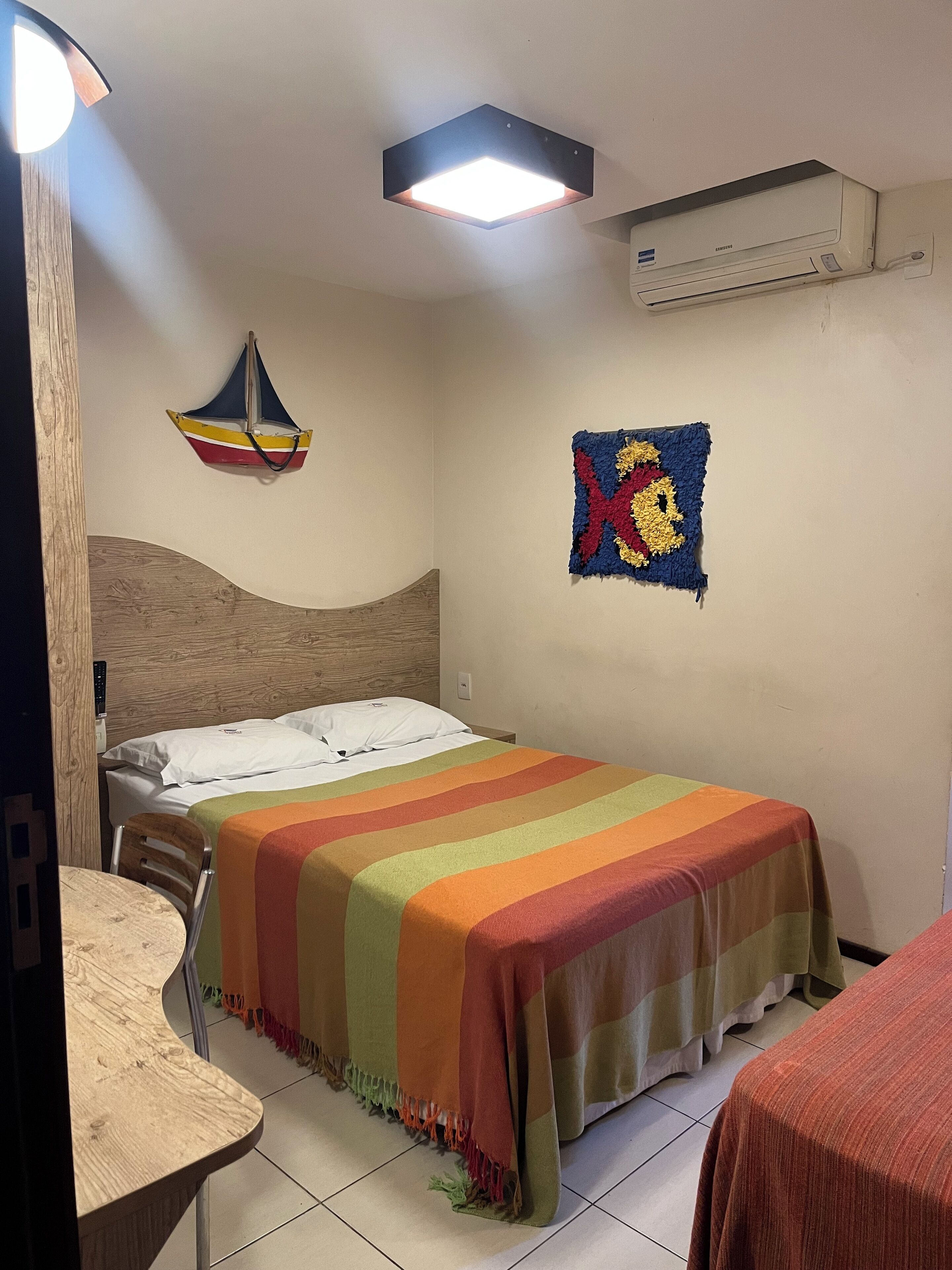 Une chambre d’hôtel équipée d’un lit, d’une table, d’une chaise, d’un climatiseur et d’une œuvre d’art accrochée au mur.