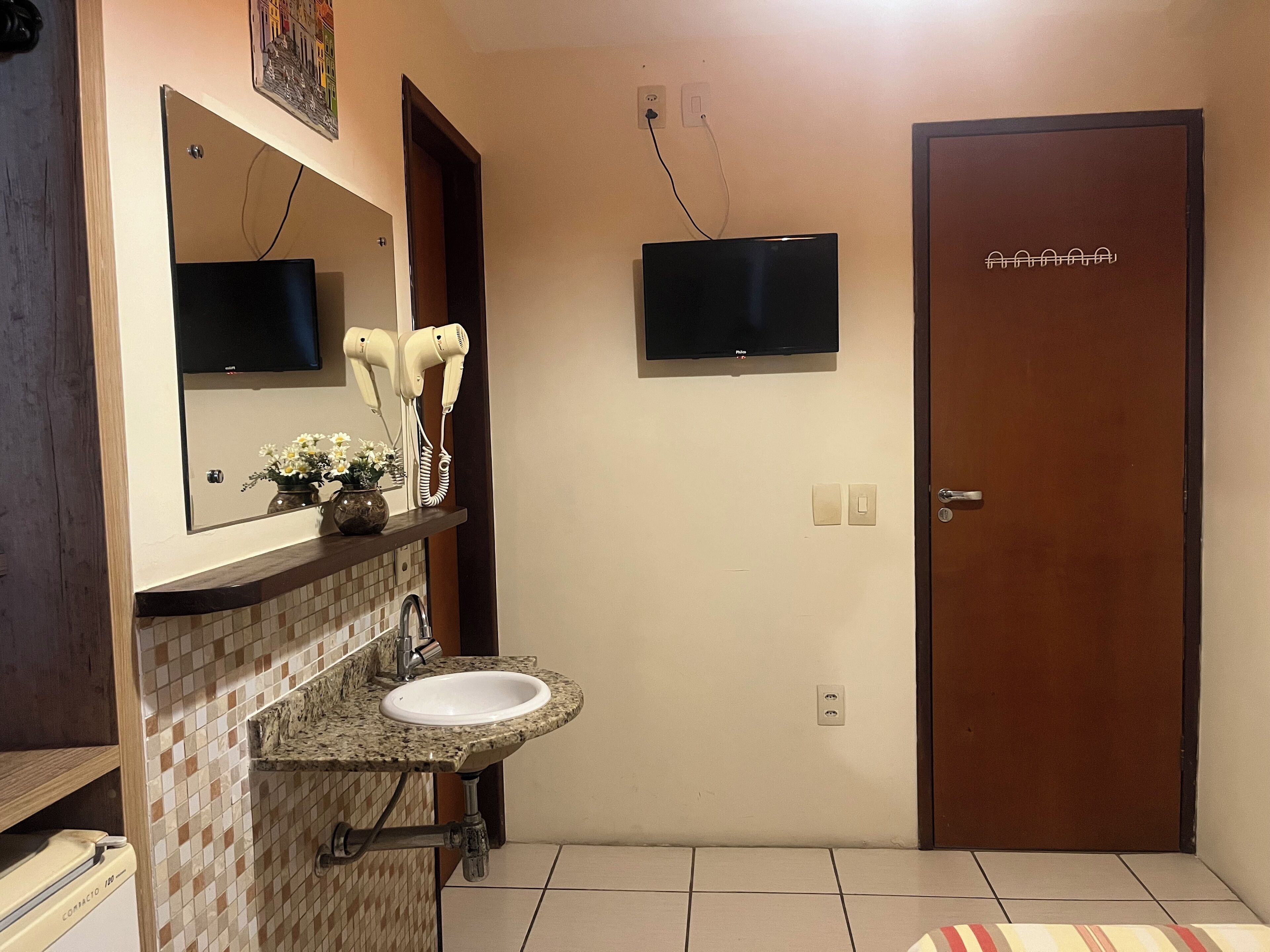 A bathroom with a sink, mirror, hair dryers, and a television on the wall.