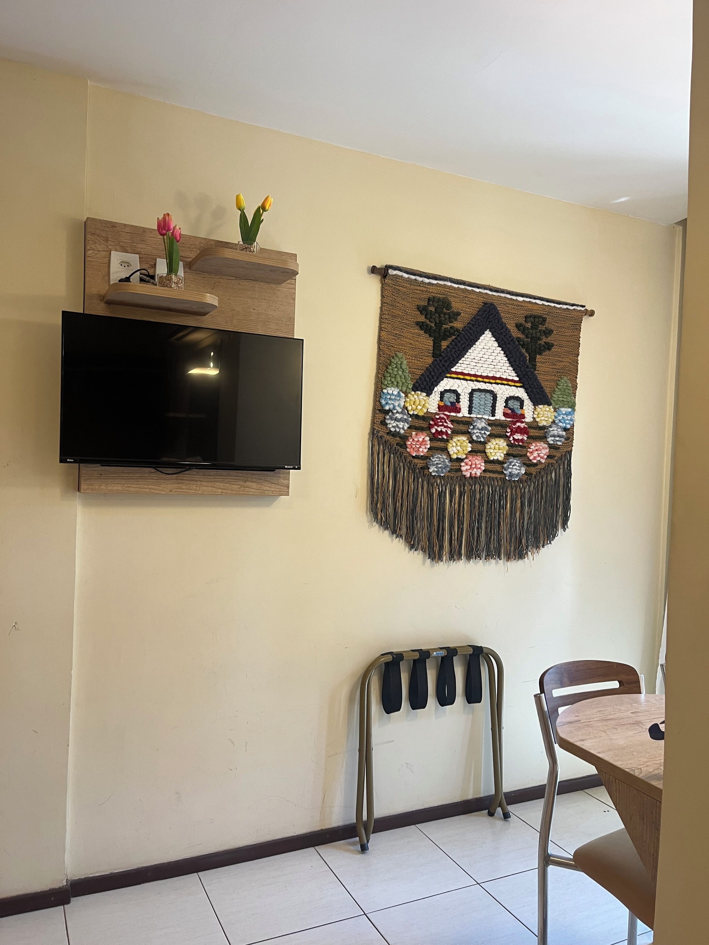 Una TV a schermo piatto montata a parete, un tappeto tessuto con un motivo di casa, e un'area pranzo con sedie in metallo e un tavolo in legno.