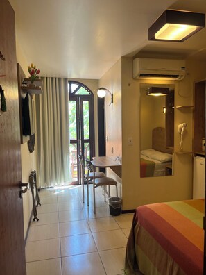 A hotel room with a bed, a desk, a chair, a small table, a trash bin, and a bathroom with a mirror and a toilet. - Vitória Praia Hotel (Recife)