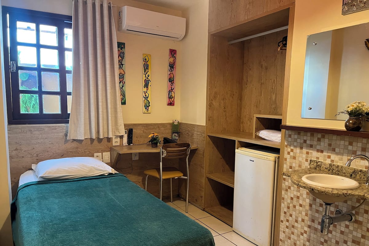 A compact hotel room with a bed, a desk, a chair, a small refrigerator, and a bathroom with a sink and mirror.